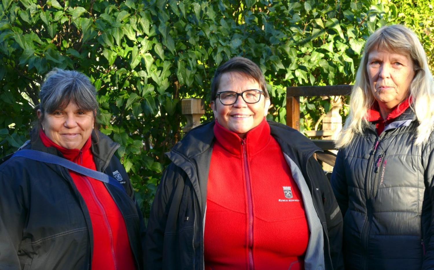 Tre personer i röda och svarta jackor står utomhus och ler, med grönska i bakgrunden.