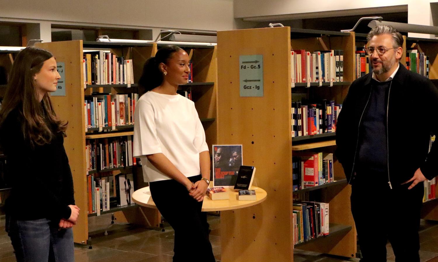 Tre personer står och pratar i ett bibliotek. Två kvinnor är nära ett runt bord med böcker, medan en man står i närheten. Hyllor fyllda med böcker finns i bakgrunden.