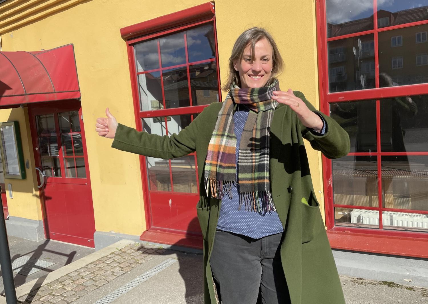 Person i grön rock och rutig halsduk står utanför en gul och röd byggnad med armarna utsträckta och ler.