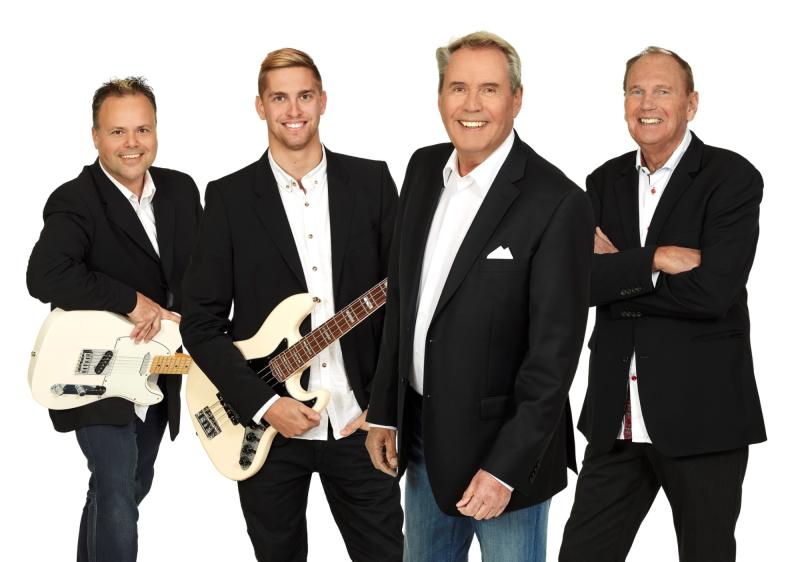 Fyra män i svarta kavajer poserar tillsammans, varav två håller i elgitarrer, mot en enfärgad vit bakgrund.