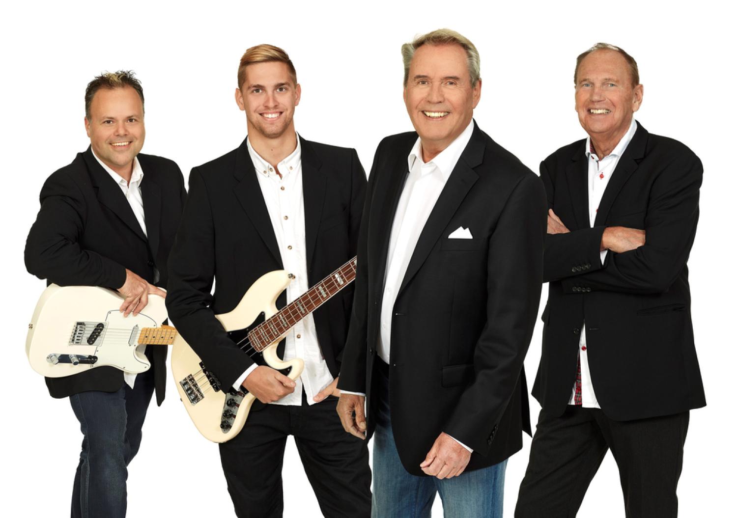 Fyra män i mörka kavajer, tre med musikinstrument och en stående med armarna i kors, poserar mot en vit bakgrund.