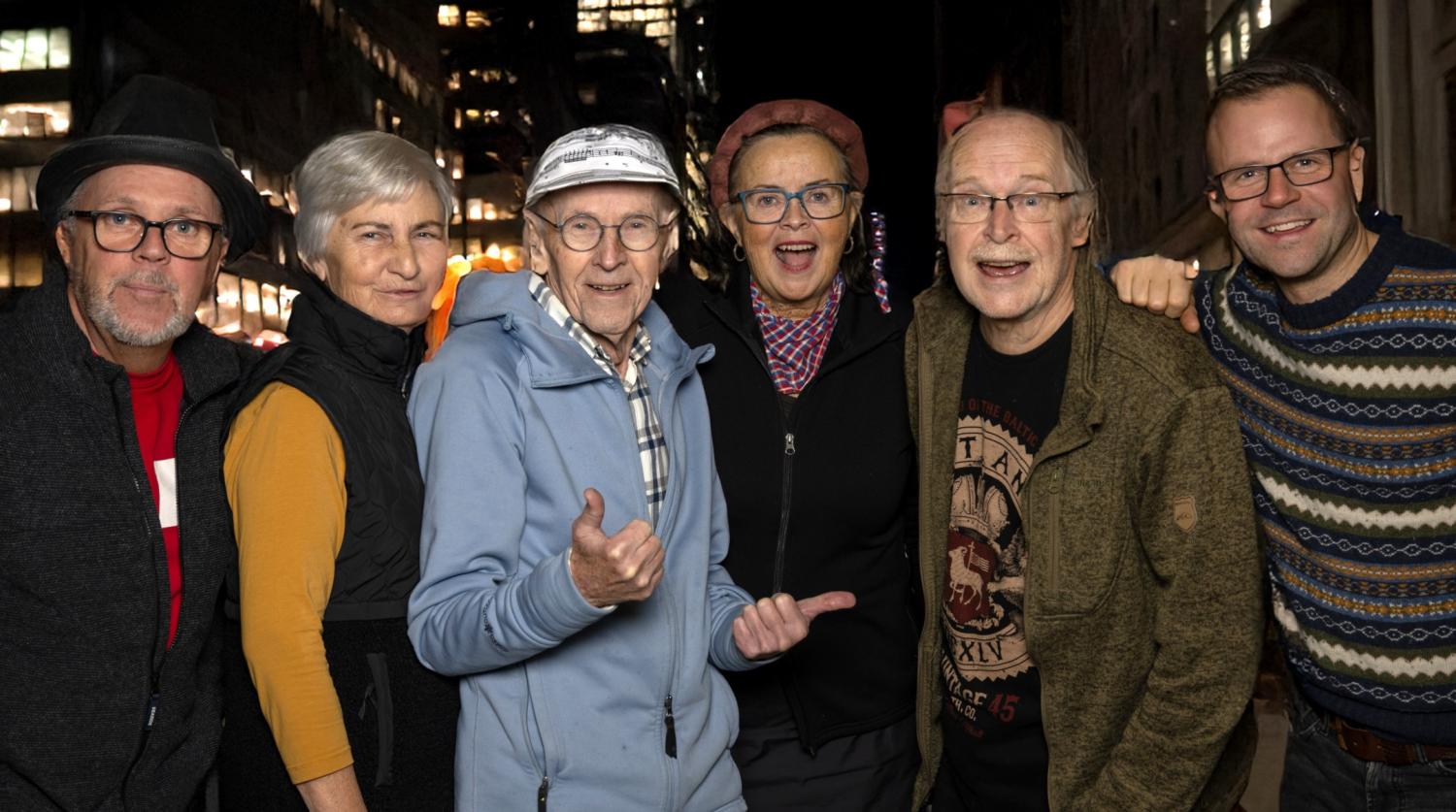 Sex vuxna står sida vid sida på natten på en gata i en stad och ler mot kameran. Några bär glasögon och hattar och i bakgrunden syns byggnader med belysning.