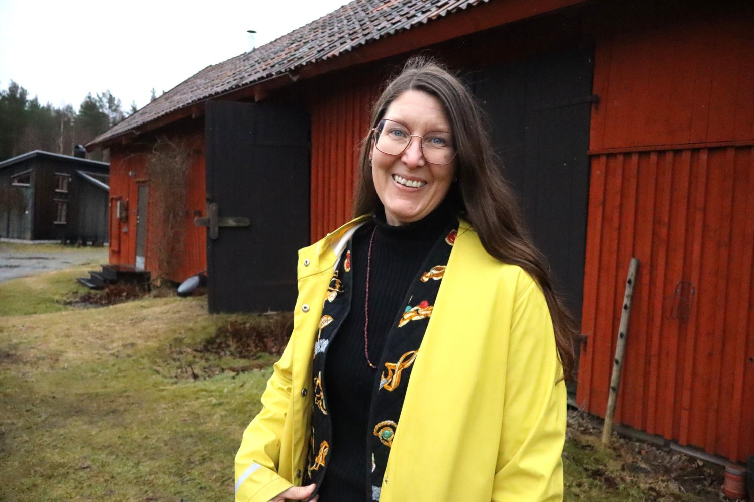 En kvinna i gul regnrock och glasögon står utanför en röd träbyggnad en molnig dag.