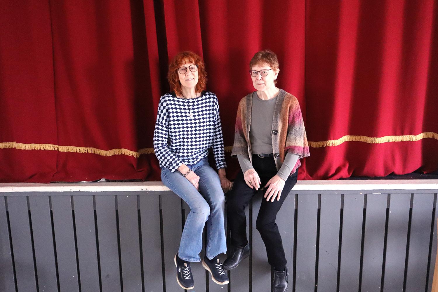 Två kvinnor sitter sida vid sida på en scenavsats framför fördragna röda gardiner.