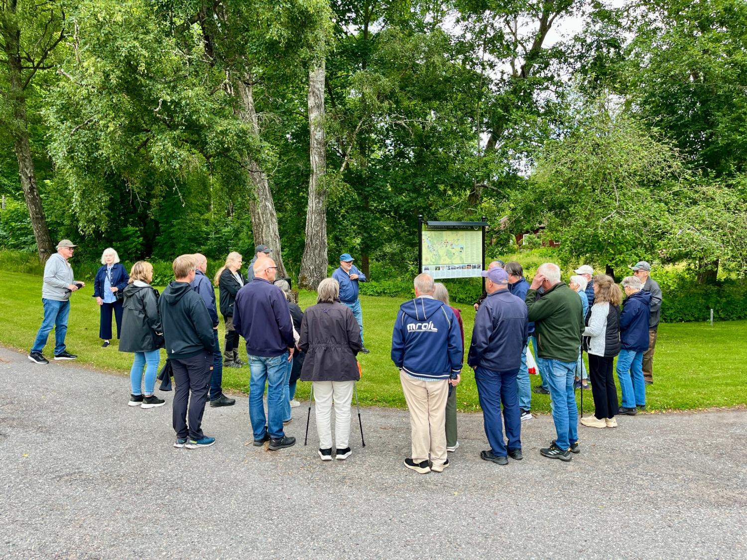 En grupp människor står på en stenlagd stig i en park, samlade runt en skylt eller karta, omgivna av gröna träd och gräs.