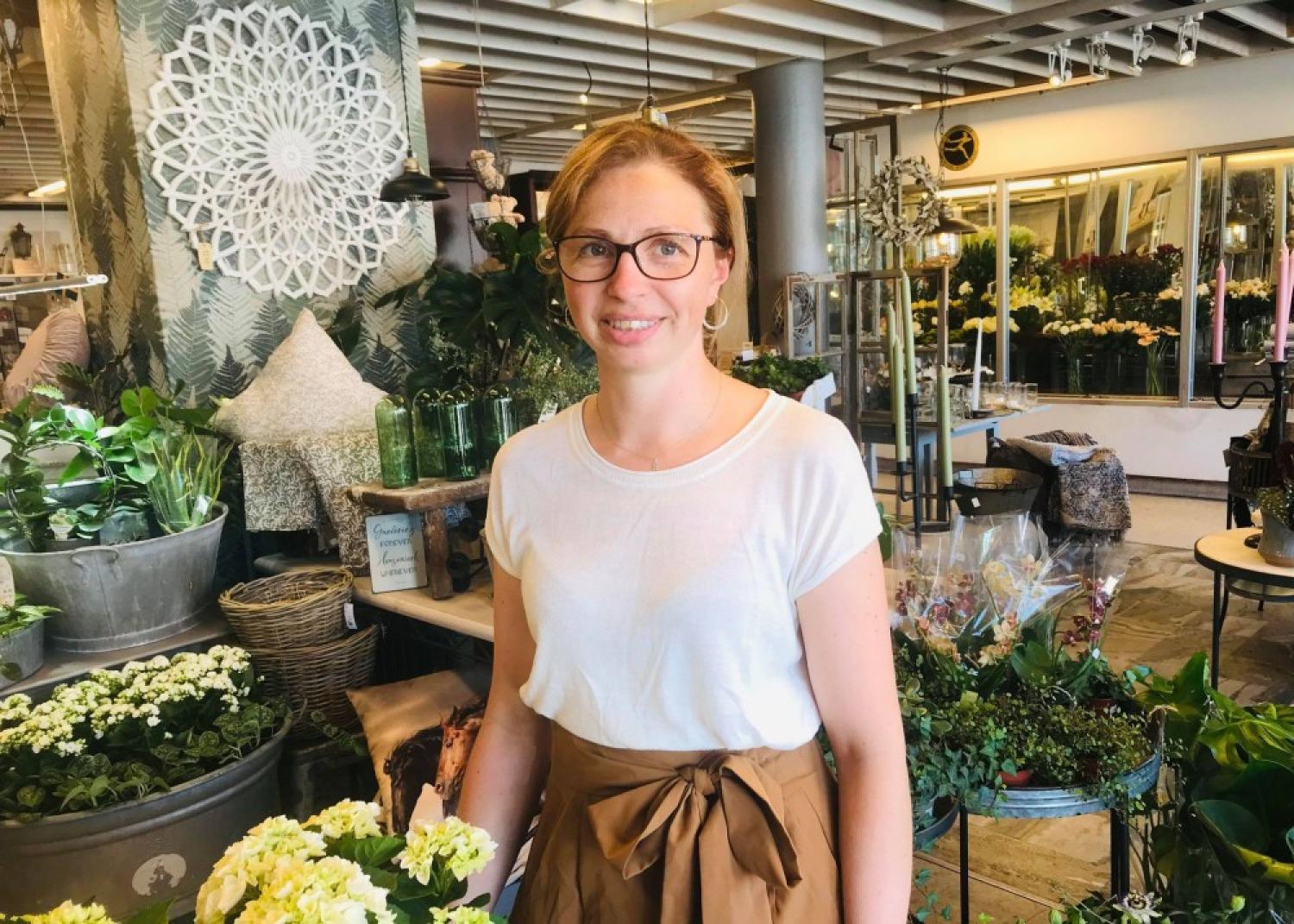 En kvinna med glasögon och vit skjorta står i en blomsteraffär omgiven av olika växter och blomsterarrangemang.