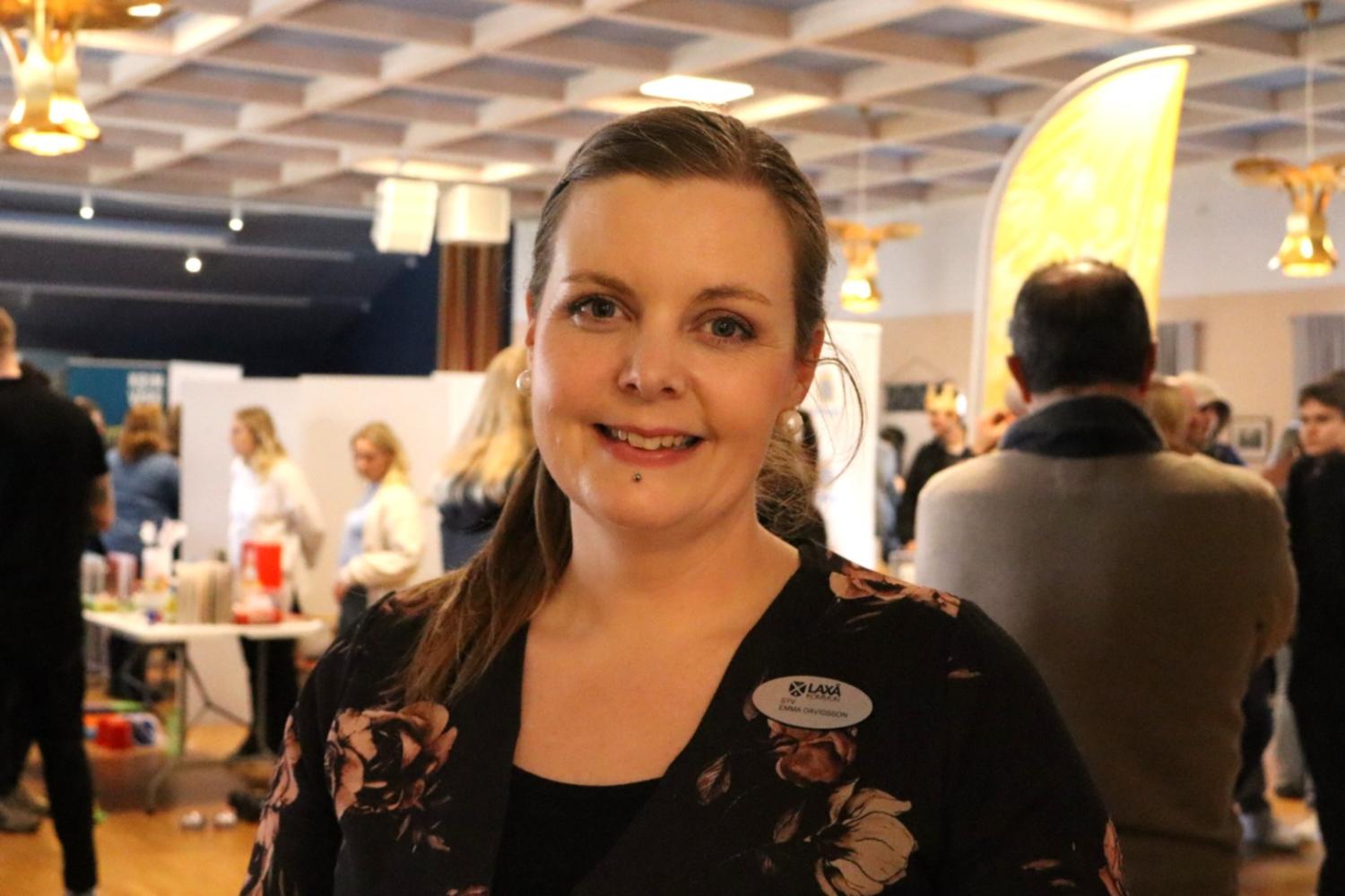 En kvinna med namnskylt ler mot kameran i en livlig inomhusmiljö med människor och bås i bakgrunden.