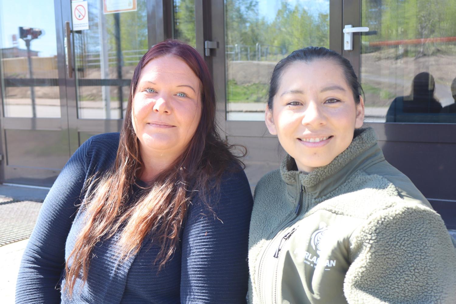 Två kvinnor sitter sida vid sida utanför en byggnad, vända mot kameran och ler i naturligt dagsljus.