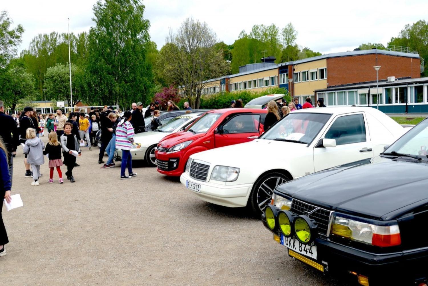 Människor samlas för att se en rad parkerade bilar, inklusive en vit sedan och ett svart fordon, i en utomhusmiljö med grönska och en byggnad i bakgrunden.