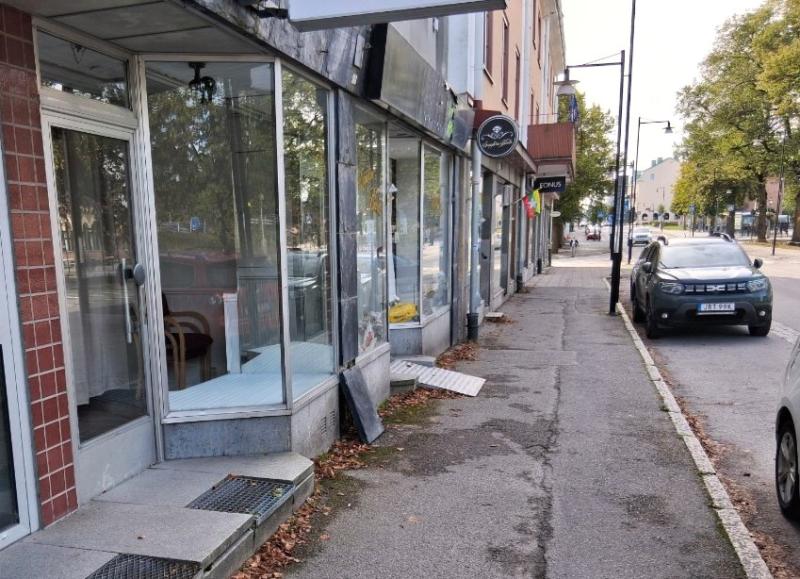 Tomma butikslokaler med glasdörrar och fönster ligger längs trottoaren på en lugn gata med parkerade bilar och några spridda löv.