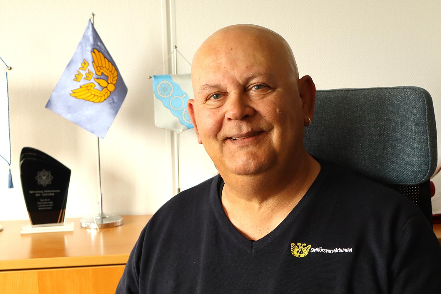 En leende skallig man sitter vid ett skrivbord med prisplaketter och blå flaggor i bakgrunden, klädd i en mörk skjorta med en broderad gul logotyp på bröstet.