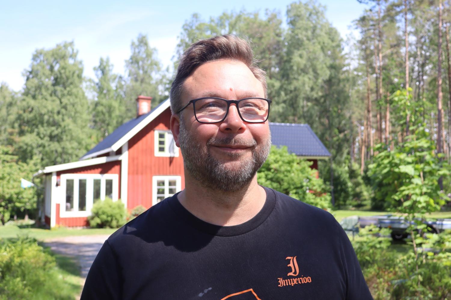 En man med glasögon och skägg står utomhus framför ett rött trähus omgivet av träd och grönska en solig dag.
