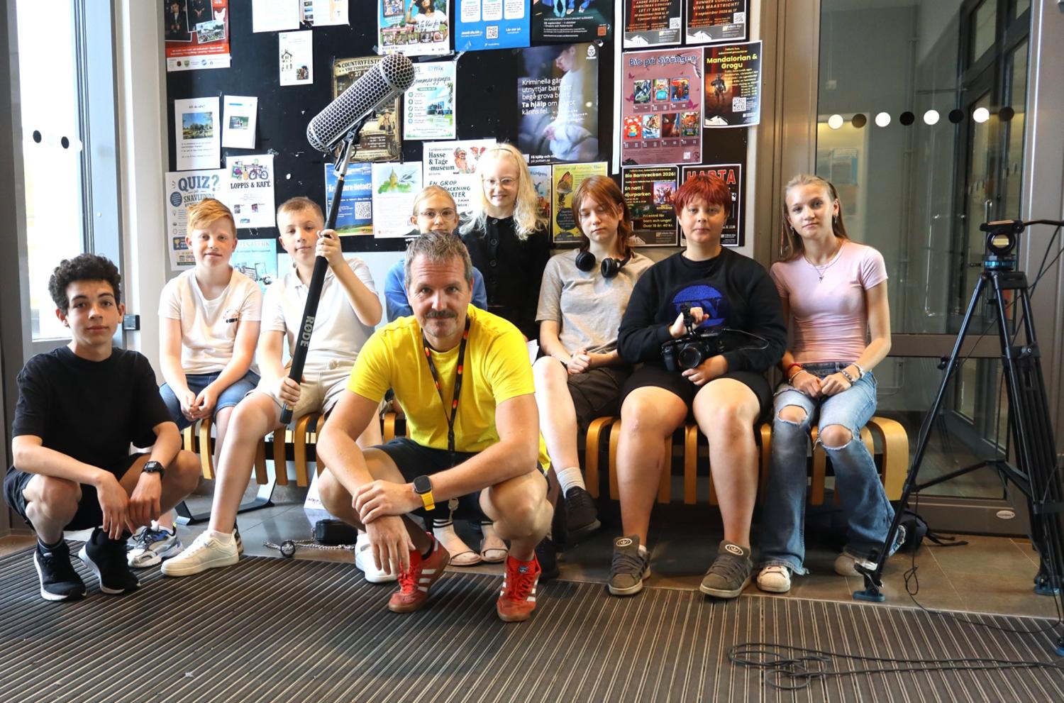 En grupp bestående av åtta ungdomar och en vuxen man poserar inomhus med filmutrustning framför en anslagstavla täckt av affischer.