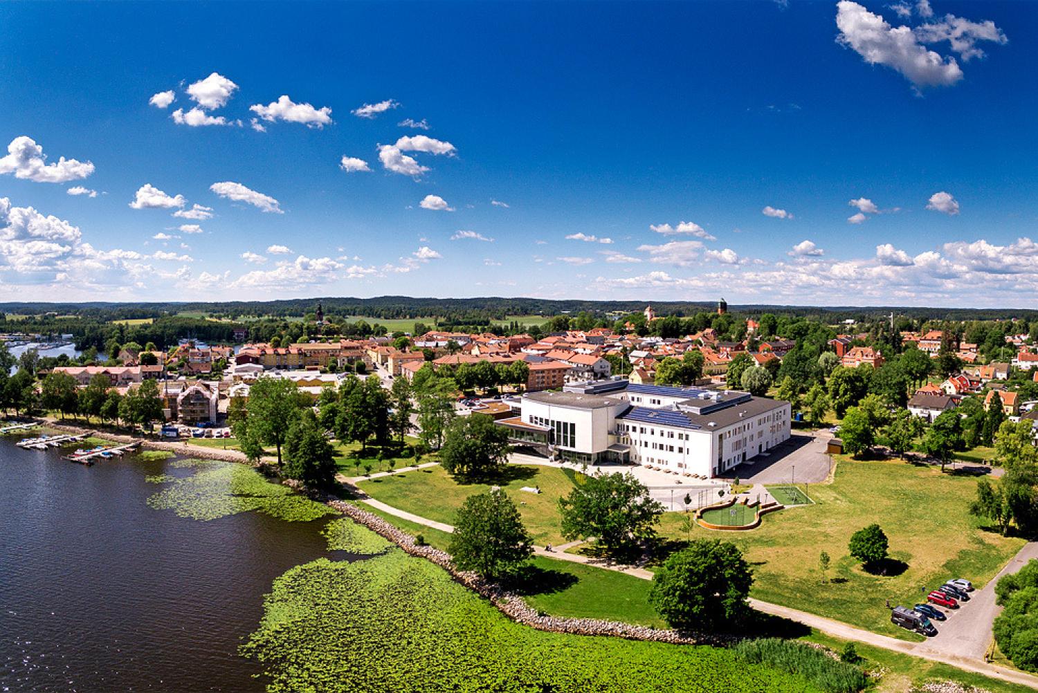 Flygfoto över en modern vit byggnad nära en flod, omgiven av gröna gräsmattor, träd och en stadsbild under en blå himmel med spridda moln.