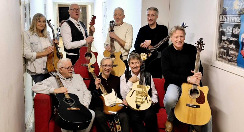 Nio personer som håller gitarrer och ukulele poserar sittande och stående runt en röd soffa i ett trångt rum.