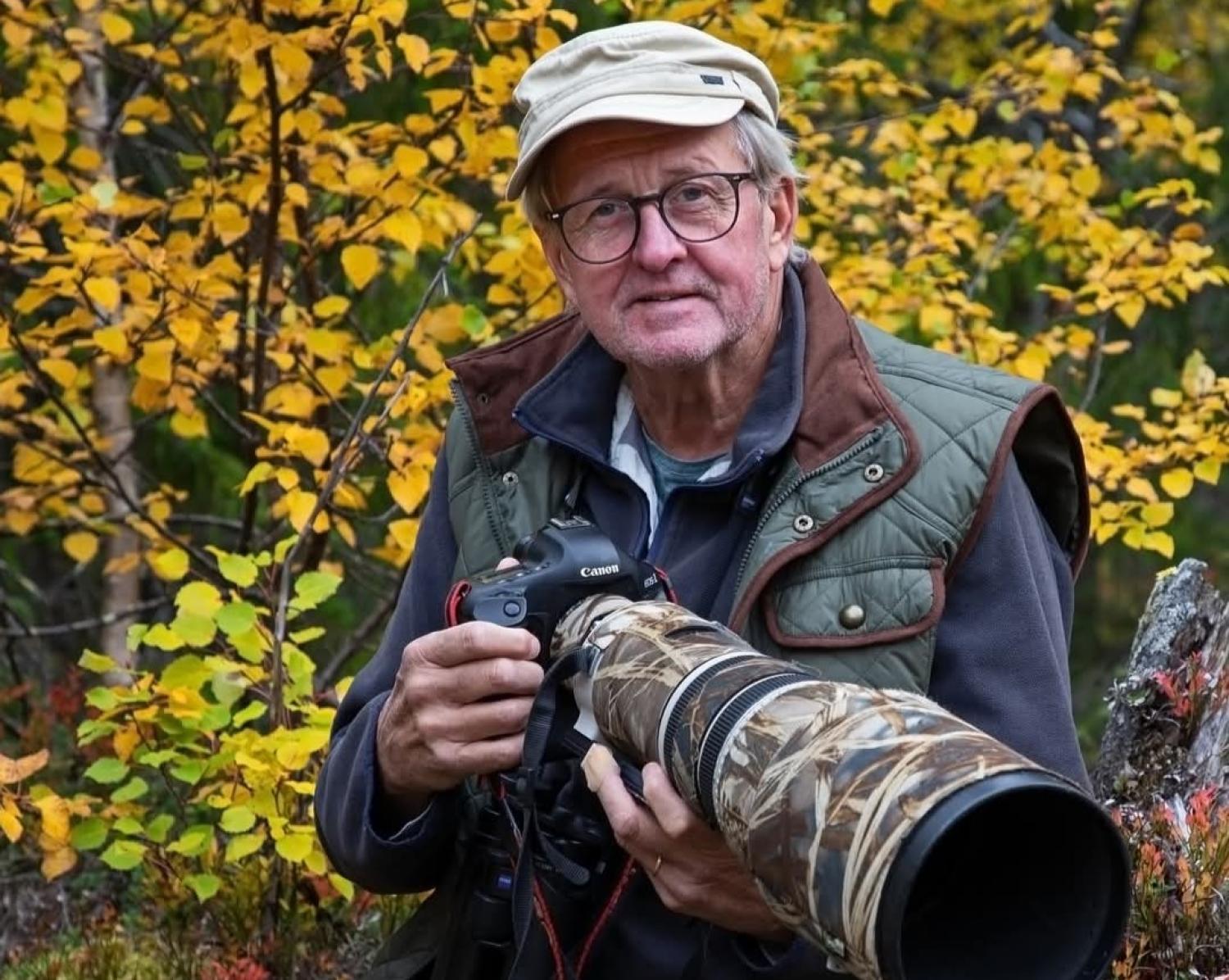 En äldre man med glasögon och friluftskläder håller i en Canon-kamera med ett stort kamouflageobjektiv och står framför höstlövverk.