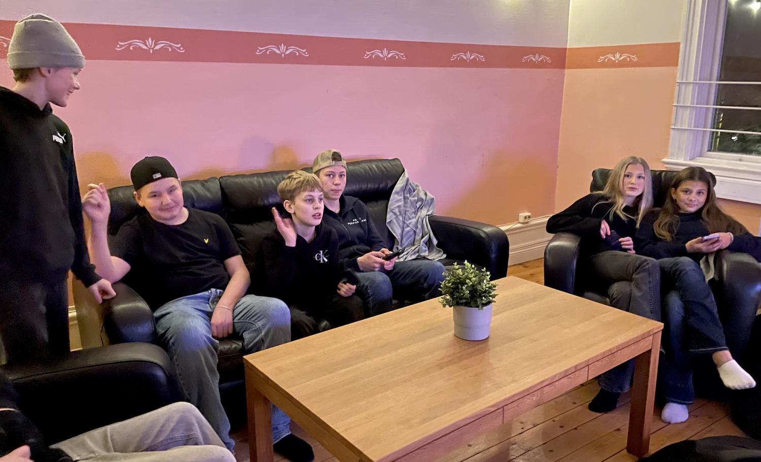 En grupp ungdomar sitter och står runt ett träbord i ett vardagsrum med en krukväxt, samtalar och använder apparater.