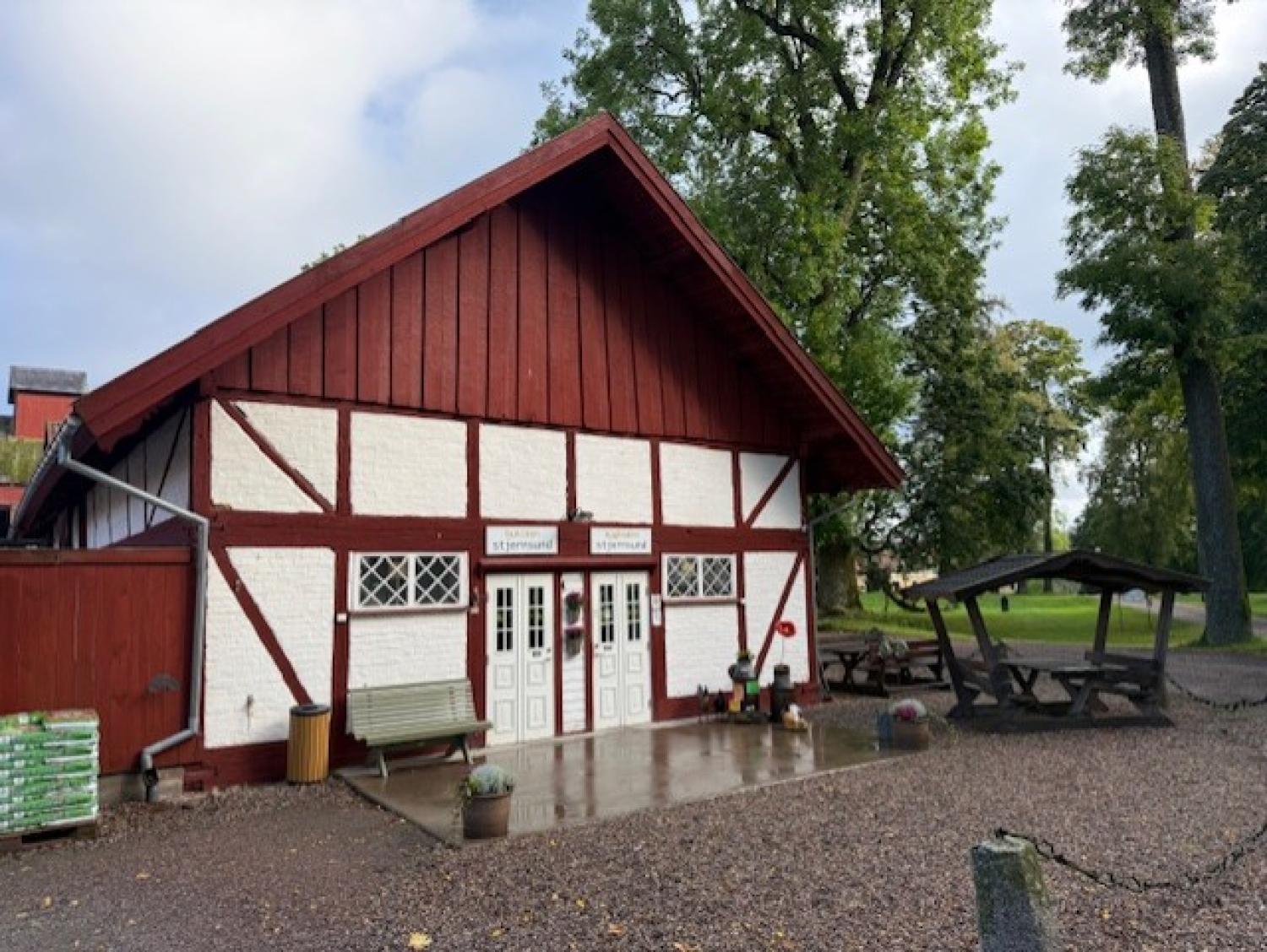 En röd och vit träbyggnad med två dubbeldörrar, utomhusbänkar, picknickbord och träd i bakgrunden. Marken är våt, vilket tyder på att det nyligen har regnat.