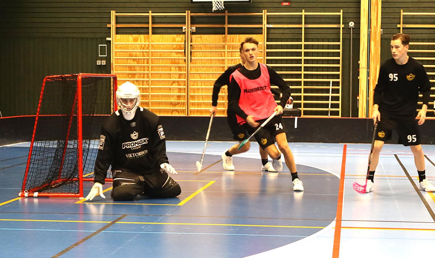 Tre innebandyspelare är i aktion nära ett nät; målvakten är knästående, medan två utespelare står och rör sig i närheten på en inomhusplan.