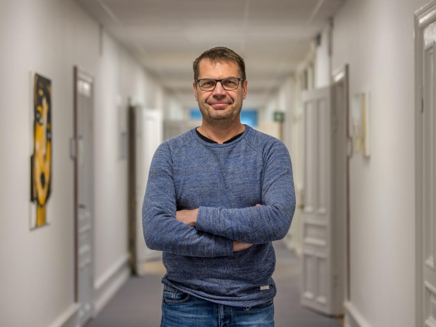 En man med glasögon står i en korridor med armarna i kors, iklädd en blå långärmad skjorta och jeans.
