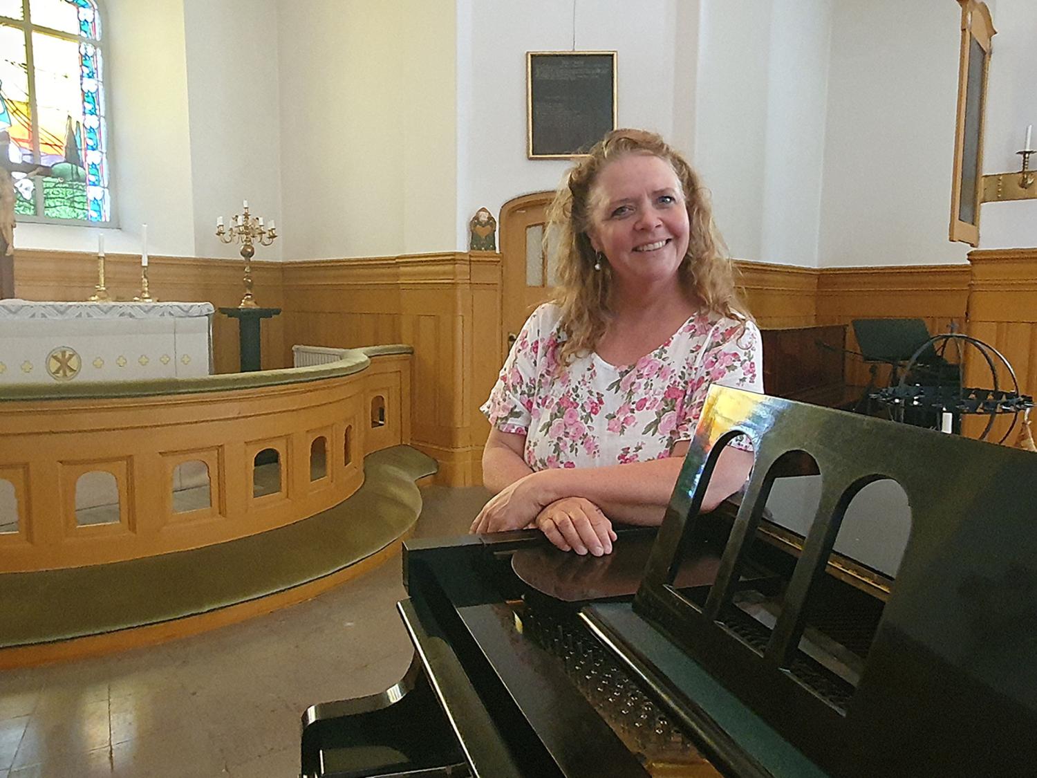 En kvinna i en blommig blus står bredvid ett piano i en kyrka med träpaneler, ett målat glasfönster och ett altare i bakgrunden.