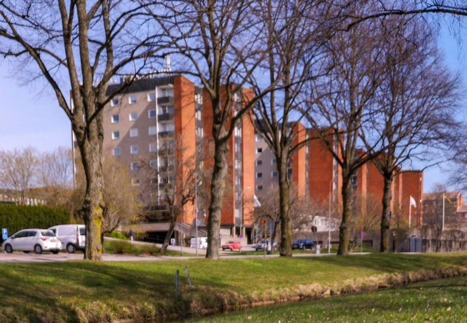 En rad hyreshus i rött tegel med balkonger skyms delvis av lövlösa träd, med parkerade bilar och en gräsyta i förgrunden.