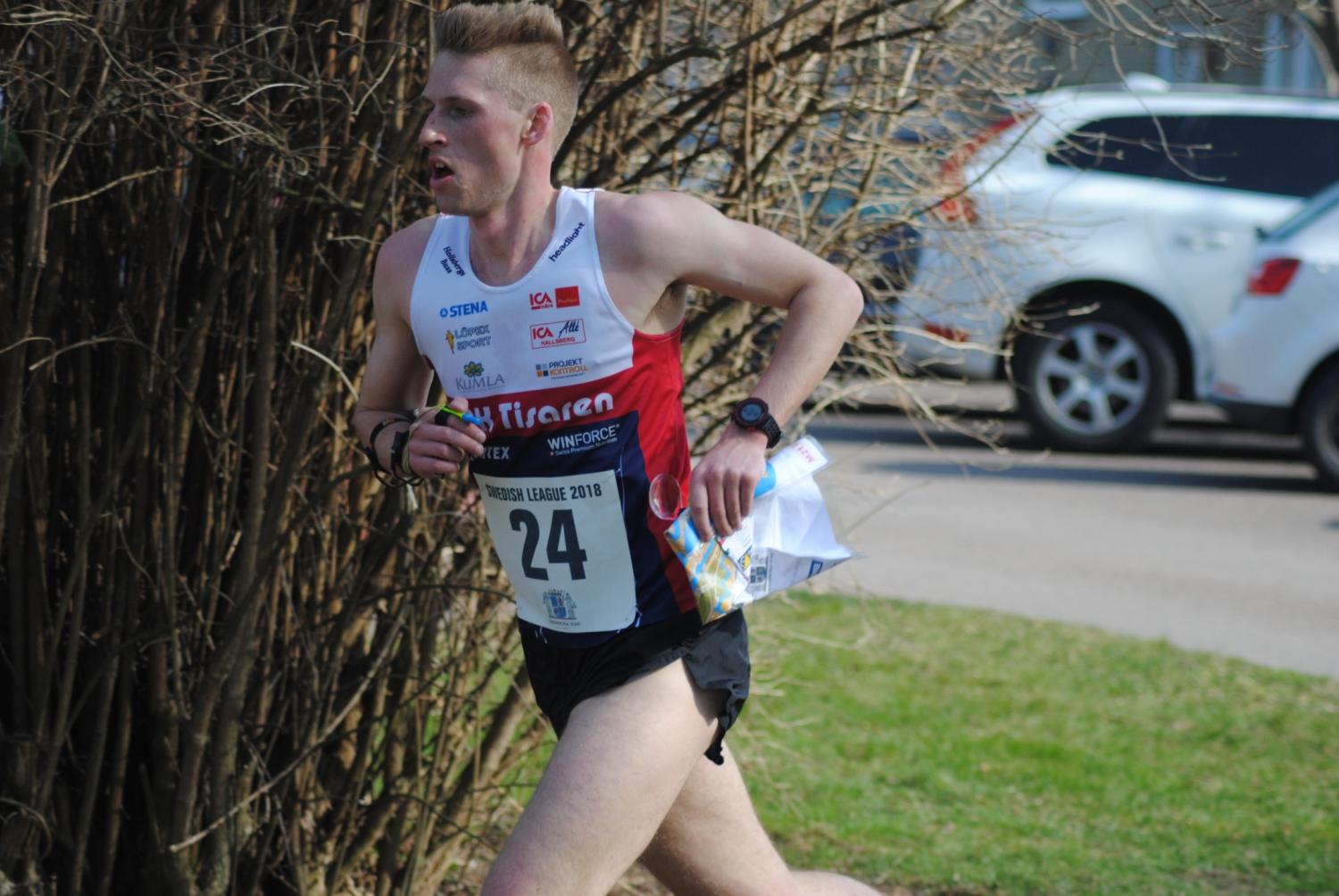 En manlig löpare med startnummer 24 och en karta i handen tävlar utomhus i närheten av buskar och parkerade bilar.
