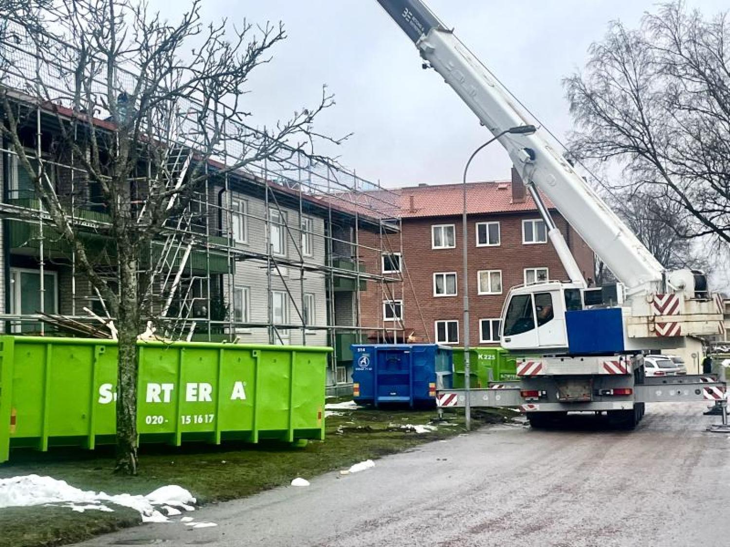 En kran lyfter en grön soptunna framför en byggnad.