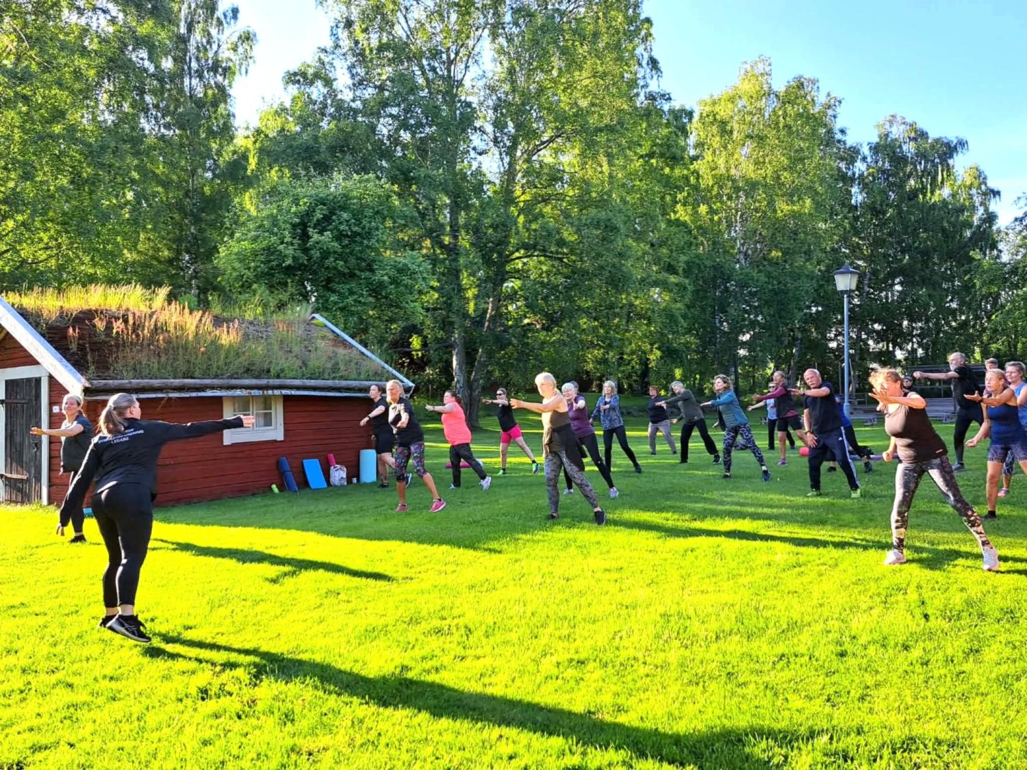 En grupp människor deltar i ett träningspass utomhus på grönt gräs nära en liten byggnad med grästak, omgiven av träd i solskenet.