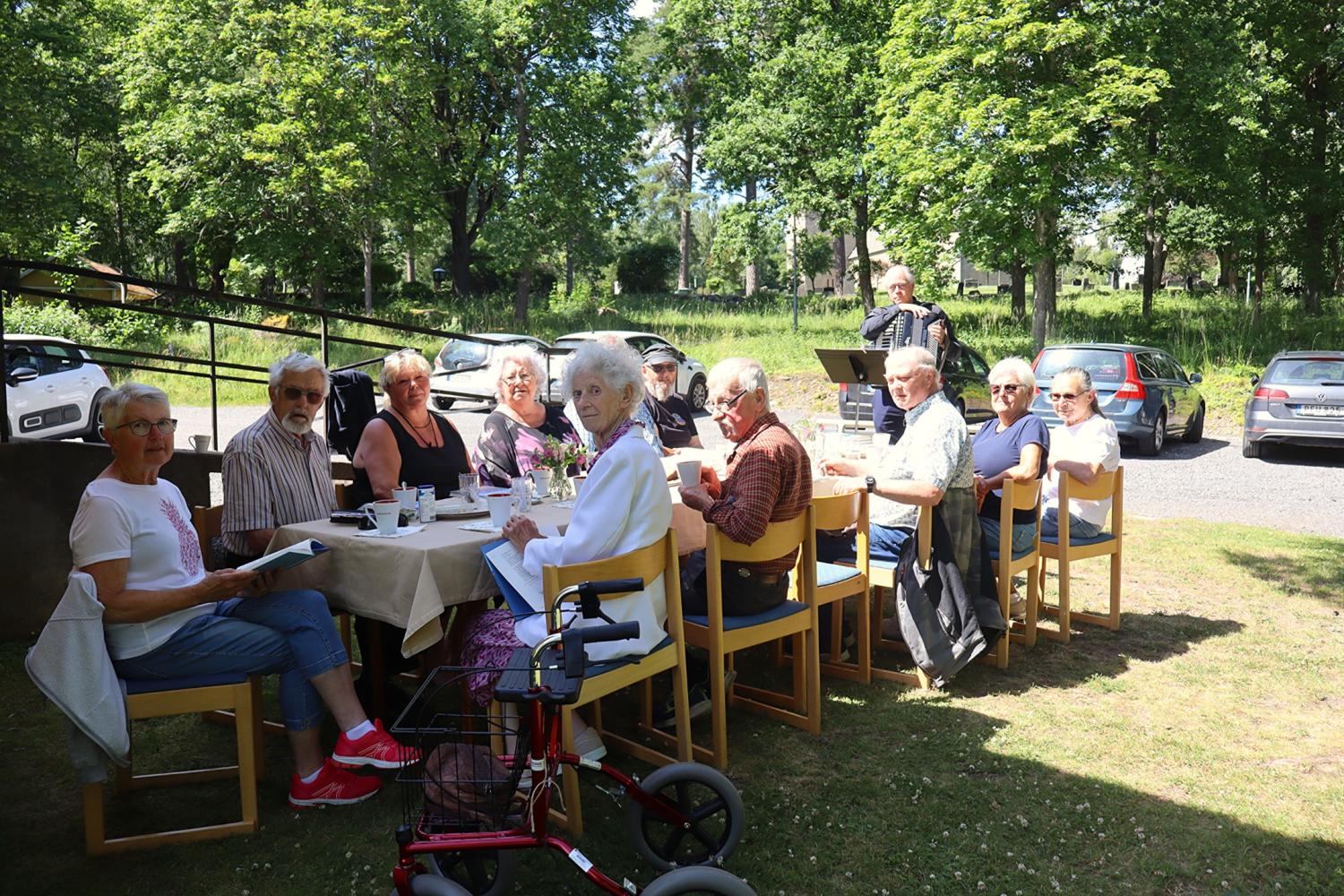En grupp äldre sitter vid ett bord utomhus och njuter av en gemensam måltid. En person i bakgrunden spelar dragspel. Bakom dem syns träd och parkerade bilar.