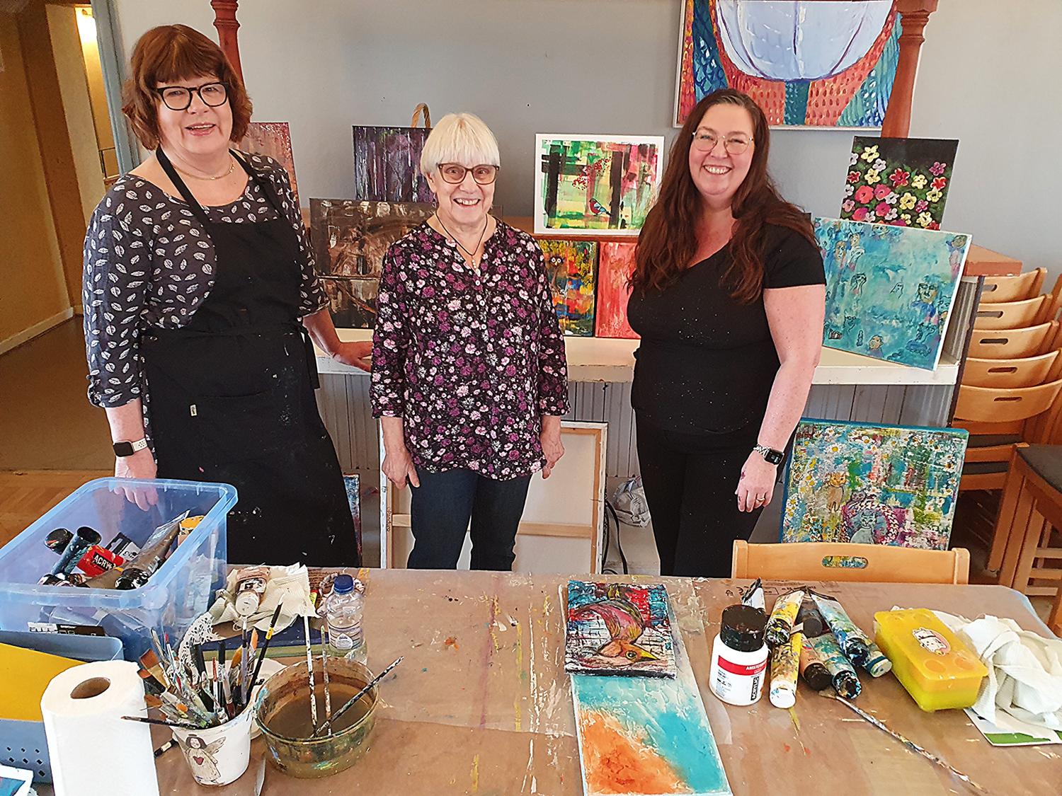 Tre kvinnor står och ler framför en utställning av färgglada målningar i en konststudio, med konsttillbehör och dukar på ett bord i förgrunden.
