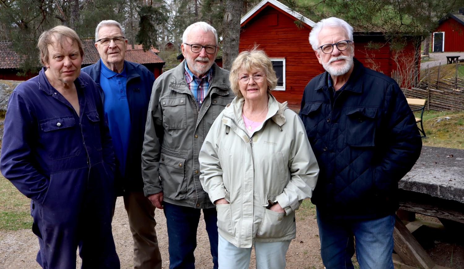Fem äldre vuxna, fyra män och en kvinna, står utomhus på en grusväg med röda träbyggnader och träd i bakgrunden.