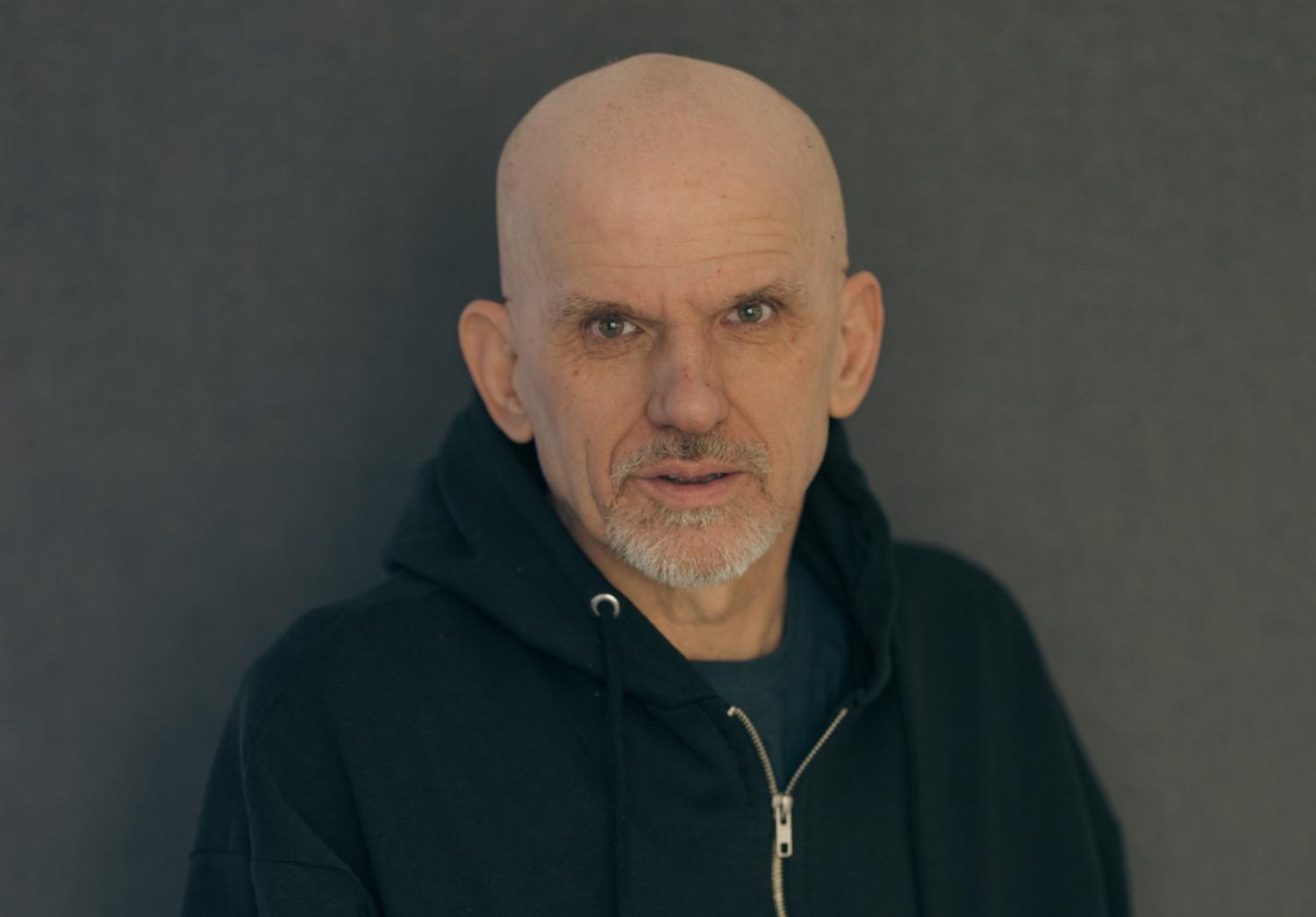 En medelålders man med skalligt huvud och grått bockskägg, klädd i svart huvtröja, står mot en grå bakgrund.