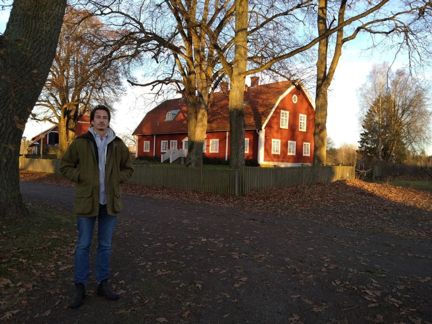 En person står på en lövbevuxen stig framför ett rött trähus med vita lister, omgiven av kala träd.