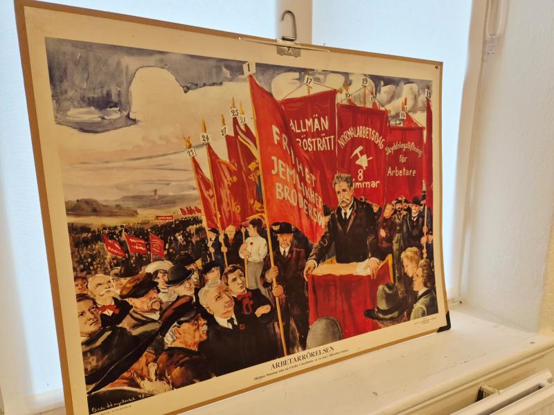 En målning föreställer en man som talar från ett podium till en folkmassa som håller röda banderoller med svensk text, som förespråkar arbetarnas rättigheter och allmän rösträtt.