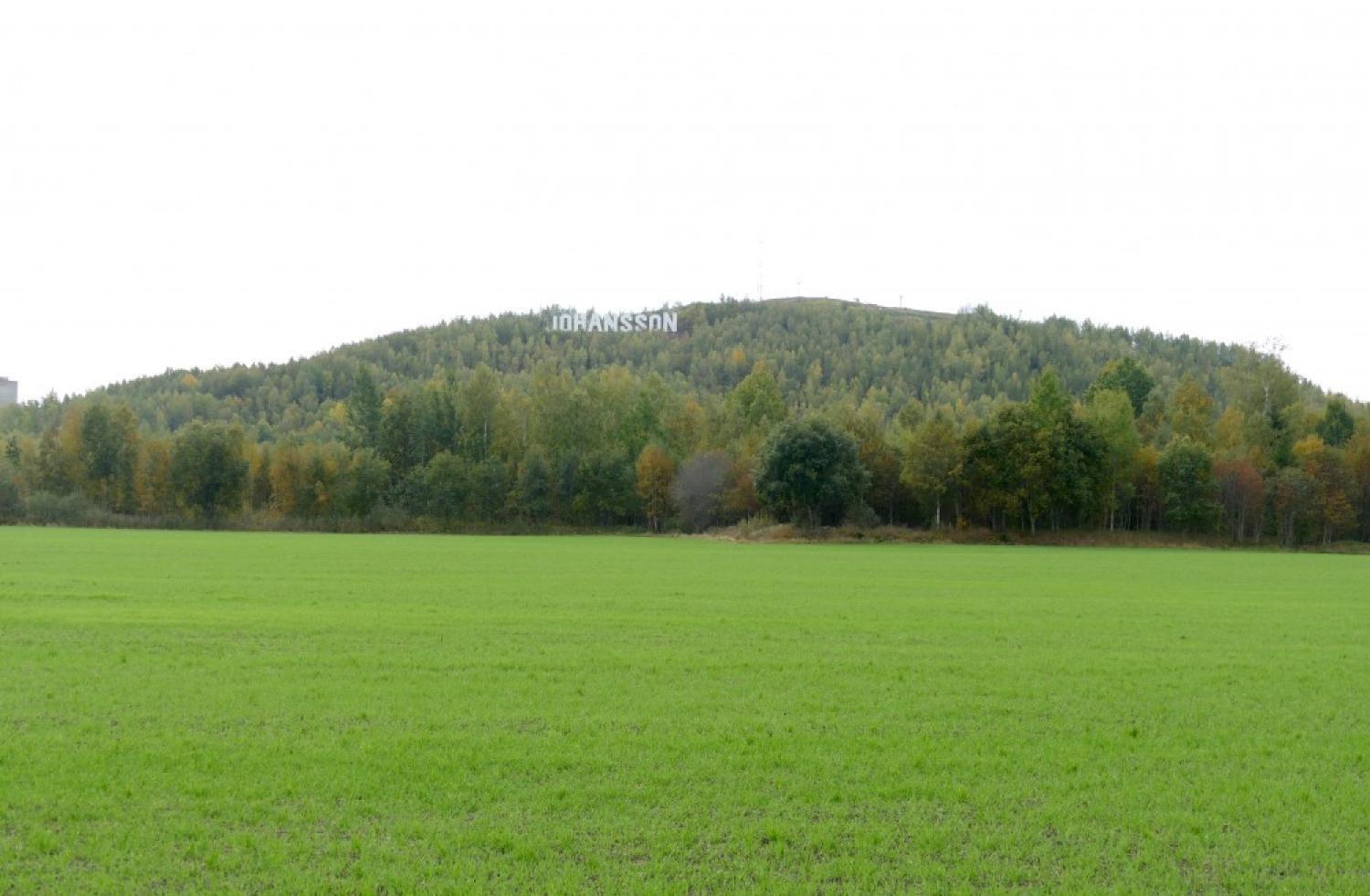 Ett brett, grönt fält som leder upp till en trädklädd kulle med några antenner på toppen under en molnig himmel.