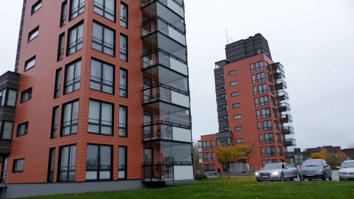 Moderna höghus med orange och svart exteriör, glasbalkonger och parkerade bilar en molnig dag.