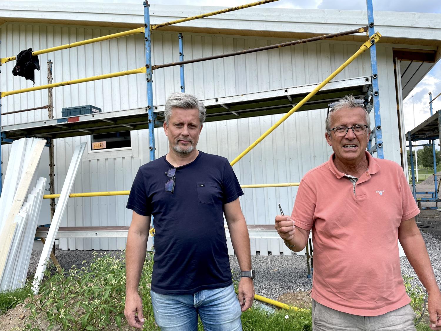 Två män står framför en vit byggnad under uppbyggnad, med byggnadsställningar och verktyg synliga i bakgrunden. En man är klädd i en mörkblå skjorta och jeans, och den andra är i en rosa pikétröja och håller i en penna.