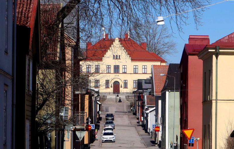 En gata kantad av parkerade bilar leder uppför en backe till en stor historisk byggnad med rött tak och årtalet 1903 inskrivet ovanför ingången.