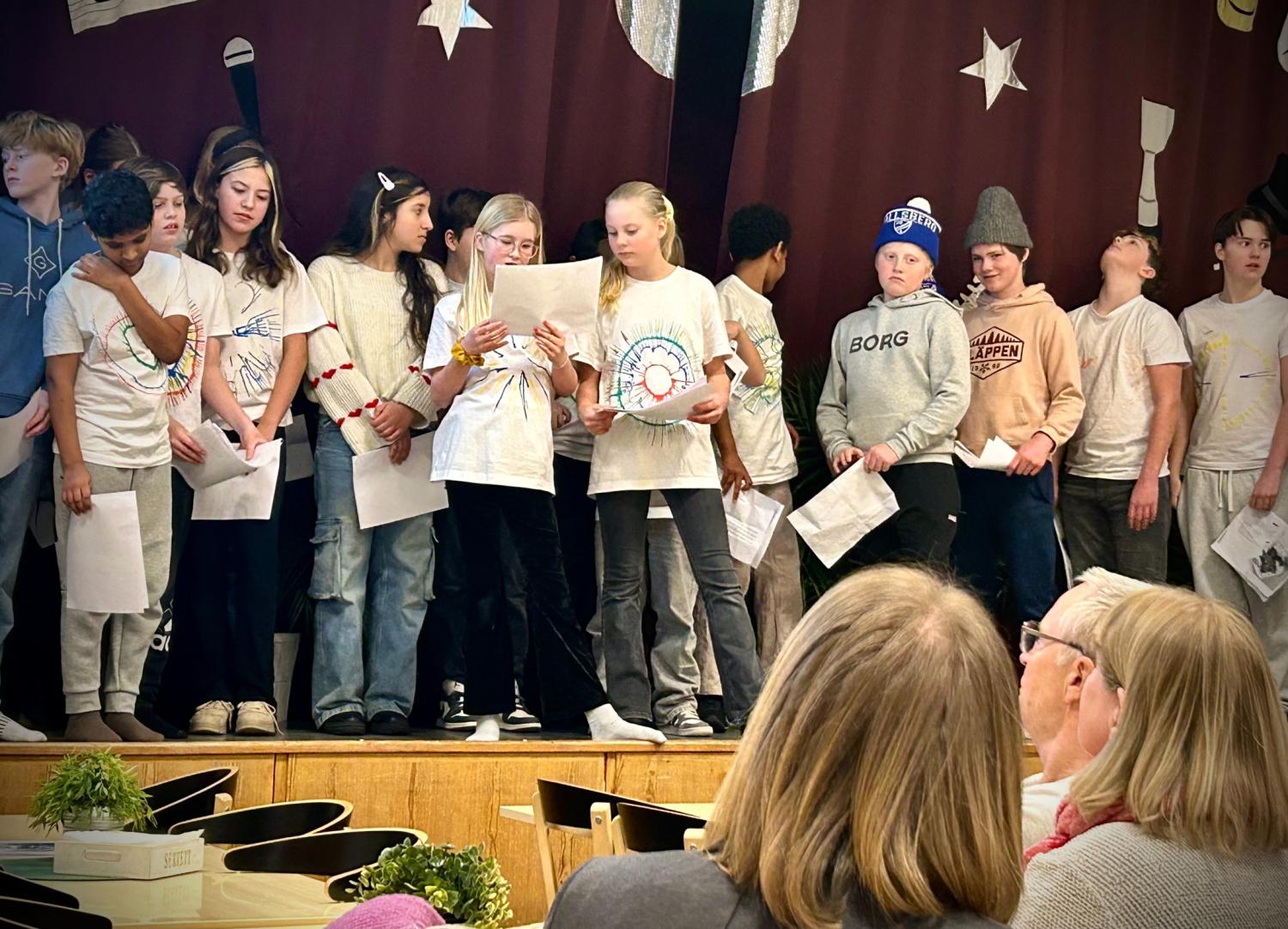 En grupp barn står på scenen med papper i handen och uppträder inför en publik som sitter vid bord med växter på. Bakom dem hänger en rödbrun gardin med dekorationer av stjärnor och penslar.