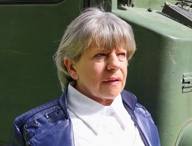 En äldre person med kort grått hår, klädd i vit skjorta och blå jacka, står i solljus framför ett grönt militärfordon.
