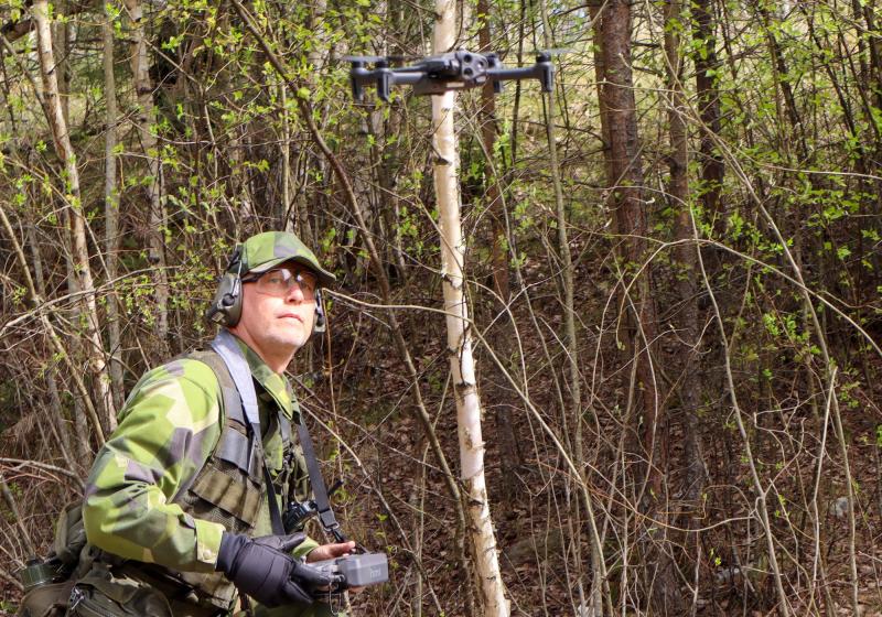 Person i kamouflageuniform som styr en drönare i ett skogsområde, håller i en fjärrkontroll och tittar på drönaren.