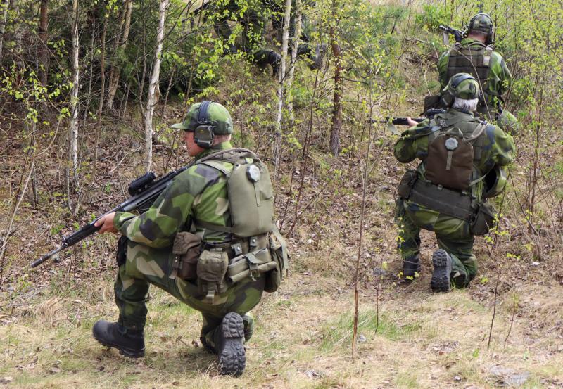 Tre soldater i kamouflageuniformer och utrustning rör sig genom ett skogsområde, hukande och siktande med sina gevär som en del av en taktisk övning.