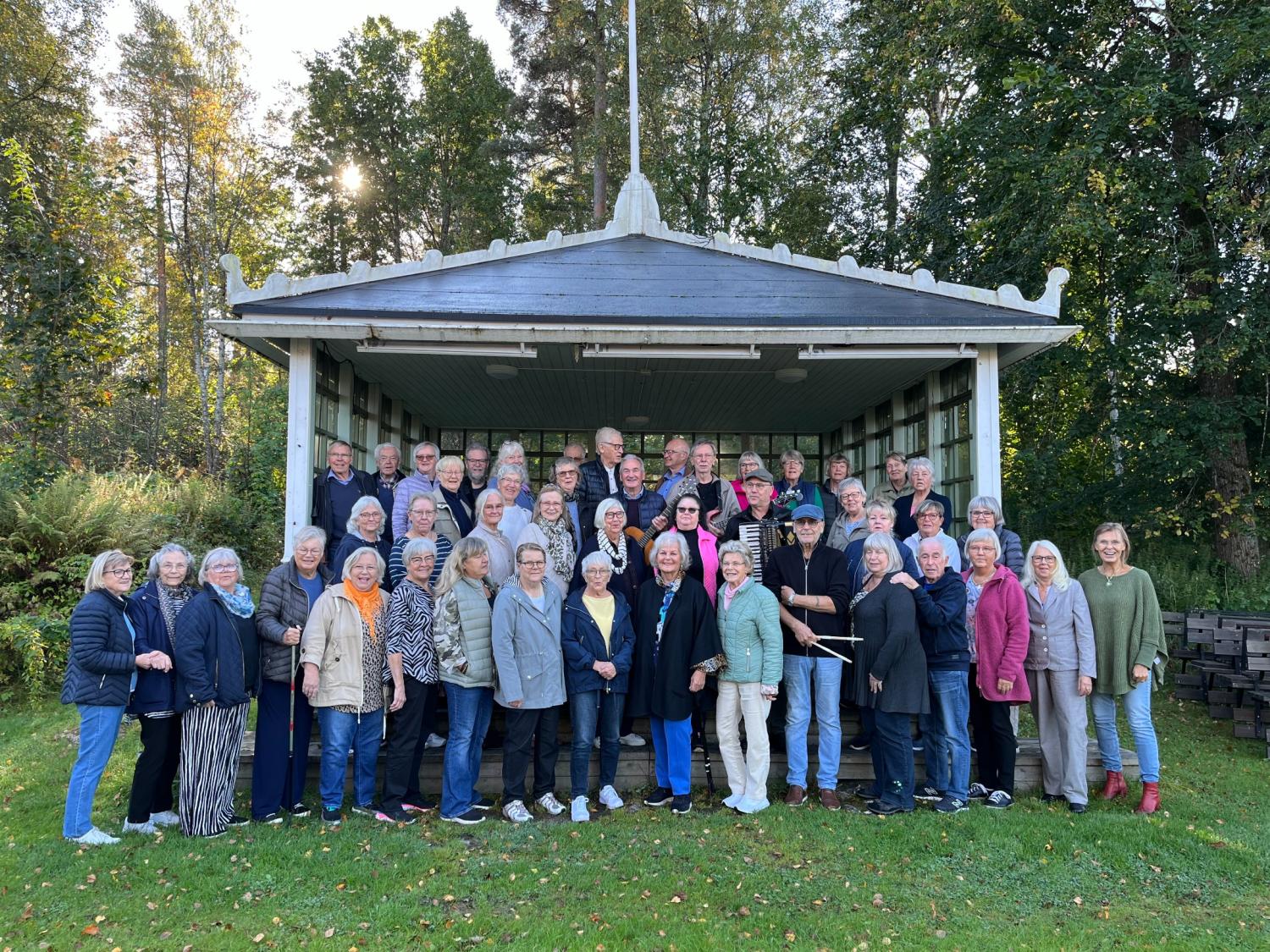 En stor grupp vuxna poserar tillsammans framför en liten utomhusscen omgiven av träd och grönska.