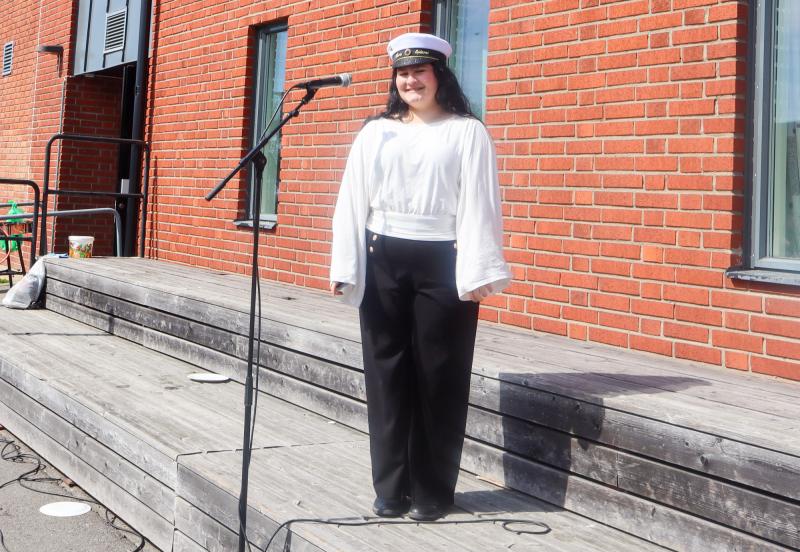 En person i vit skjorta, svarta byxor och vit hatt står framför en mikrofon på en träplattform vid en tegelbyggnad.