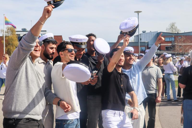 En grupp unga människor i vita studentmössor ler och lyfter på hatten utomhus under en examensfest.