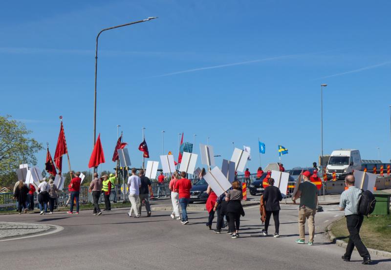 En grupp människor med tomma skyltar och flaggor har samlats på en väg nära trafikbarriärer under en klarblå himmel.