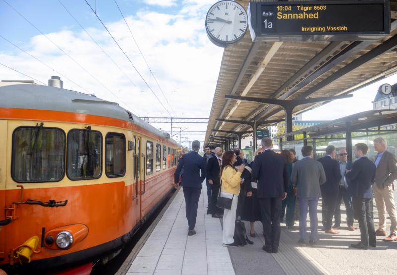 En grupp människor står på en tågperrong nära ett orange och gräddfärgat tåg, med en digital skylt som visar avgångsinformation och en stor klocka ovanför.