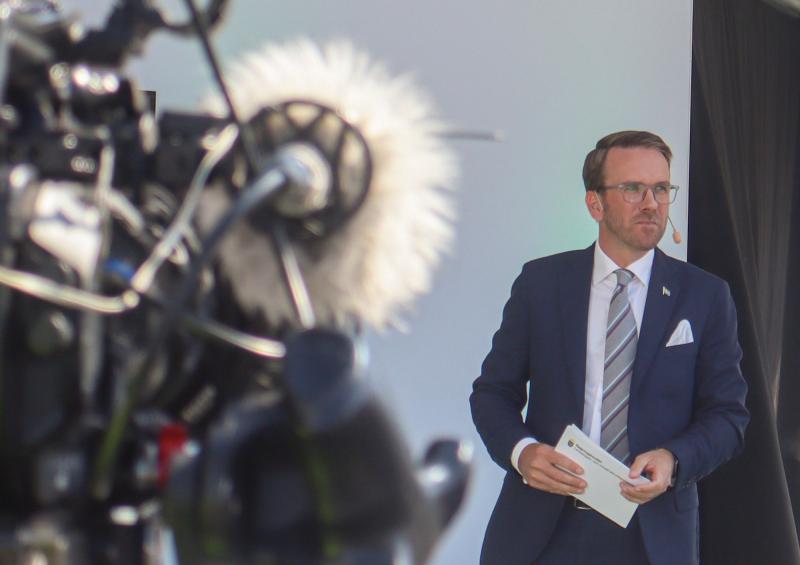 En kostymklädd man står med papper framför en professionell videokamera med headsetmikrofon och en vanlig bakgrund.