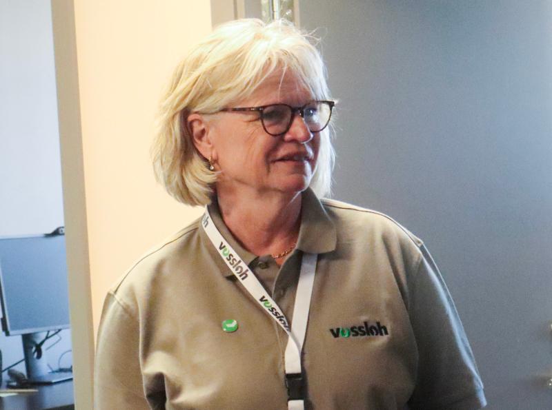 En äldre kvinna med blont hår och glasögon, klädd i khakifärgad Vossloh-polo och nyckelband, står inomhus vid en dator.